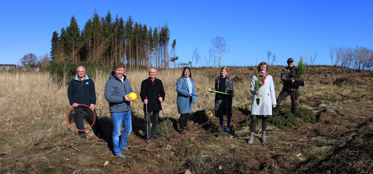 Eine Gruppe von Menschen steht im Wald auf einer Lichtung und halten Werkzeug sowie kleine Setzlinge in den Händen.